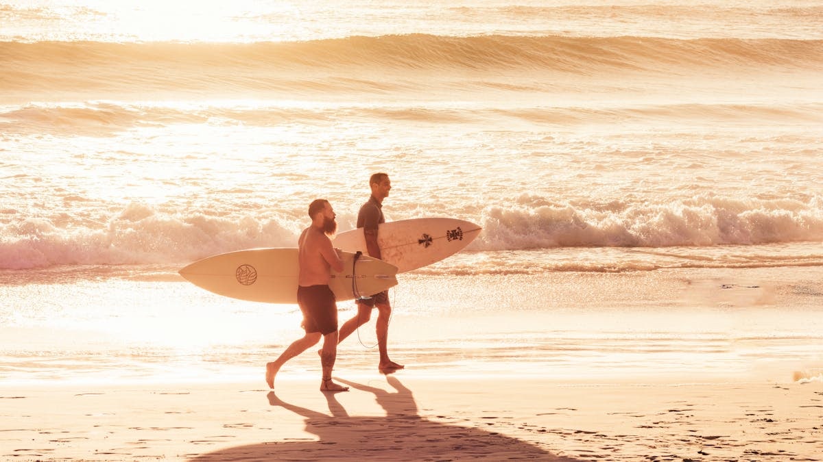Two surfers carry their boards along the sandy shore during a vibrant sunset in Arraial do Cabo.